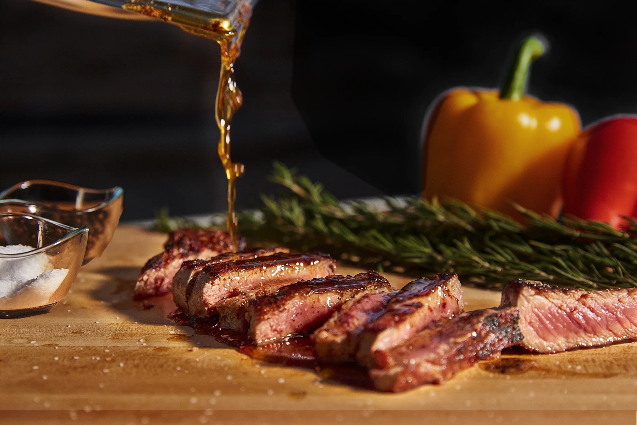 Sliced steak on a wooden cutting board with herbs and peppers, accompanied by a glass container of oil.