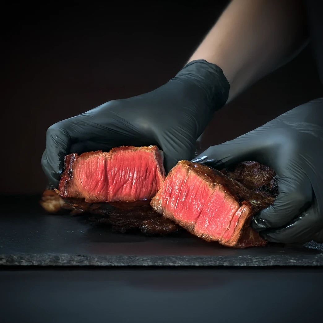 Person wearing black gloves holding a sliced piece of rare steak on a dark surface