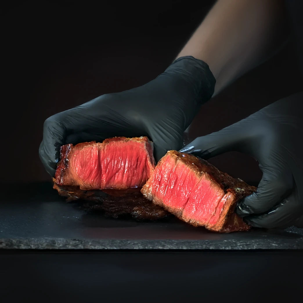 Person wearing black gloves holding a sliced piece of cooked steak on a dark surface